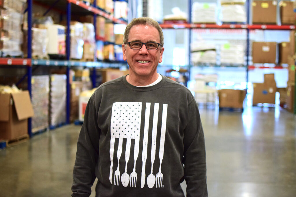 Chris Whitney, founder of OneGenAway, stands in OneGenAway's warehouse, which is full of pallets of cans and packaged food items.