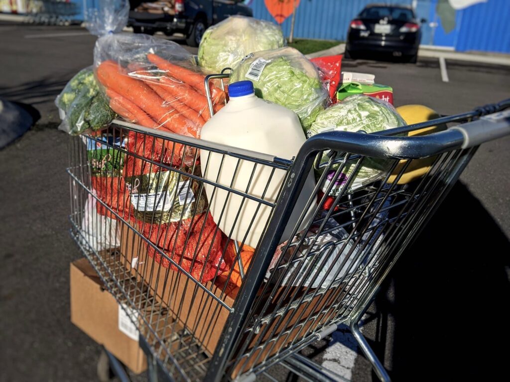 A shopping cart full of groceries including bagged and boxed items, fresh produce, and milk, stands in a parking lot at OneGenAway’s mobile food pantry.