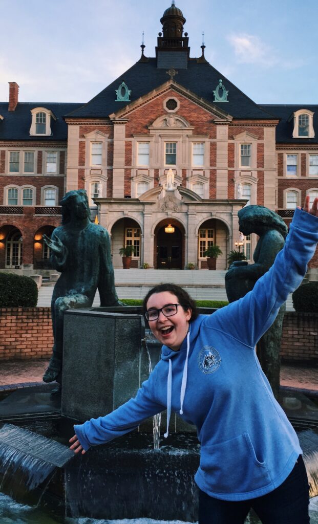 Adrienne Pelletier, a woman in a blue hoodie, strikes a pose in front of the Notre Dame Seminary building.