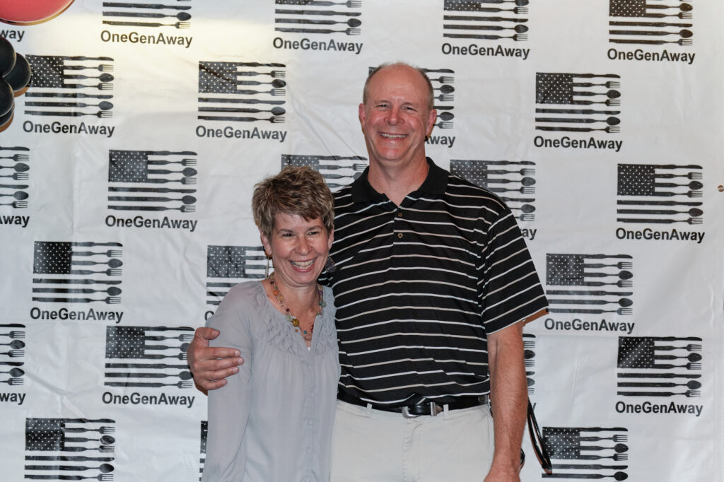 Connie Bond and Mark Bond hug on a red carpet in front of a OneGenAway logo step-and-repeat background