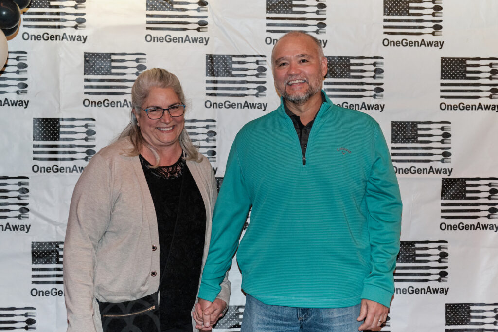 Kathy and Bobby Worthington hold hands on a red carpet in front of a OneGenAway logo step-and-repeat background