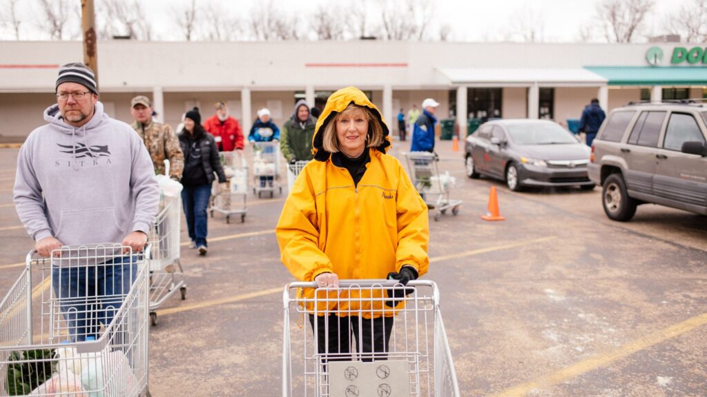 A woman in a yellow rain coat leads a line of people pushing shopping carts at the OneGenAway Mobile Pantry