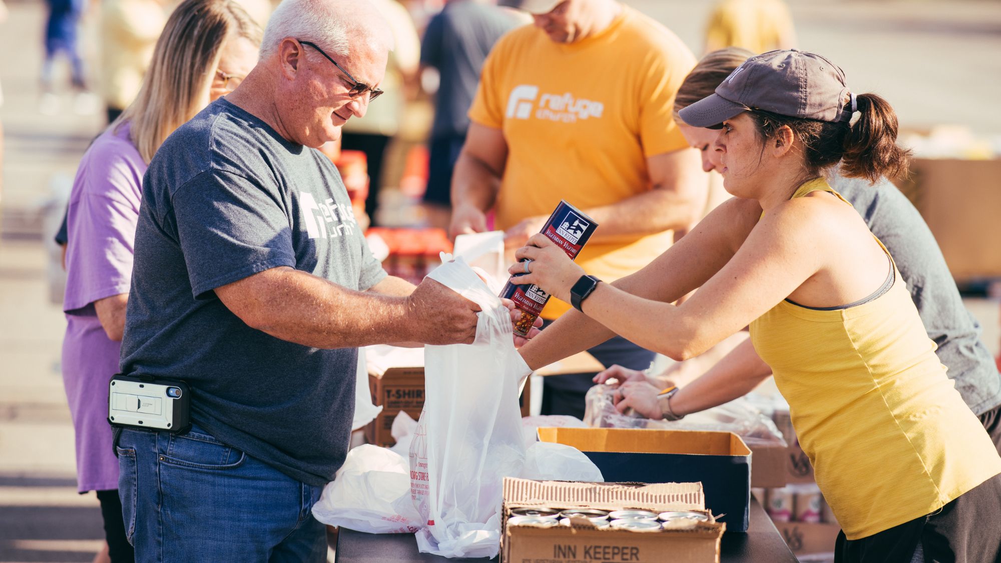 A woman in a yellow tank top and gray ball cap places canned foods into a grocery bag held by a man in a gray T-shirt