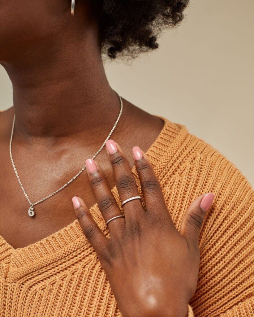 A woman in an orange knit sweater wears two silver rings on her fingers, a silver necklace with a small charm of the number 6, and silver hoop earrings.